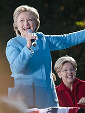 Photograph of Clinton in a light blue suit, holding a microphone and speaking in front of Elizabeth Warren who is seated behind her