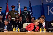 Obama whispering to Clinton at a summit meeting, with multiple soldiers in uniform standing behind them and heads of state sitting behind and in front of them. They are in the 21st NATO summit, which was held in April 2009.